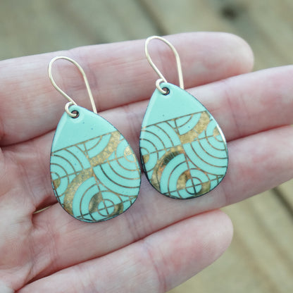 Hand holding a pair of Teardrop shaped, seafoam green enamel earrings with gold geometric designs, against a wooden background