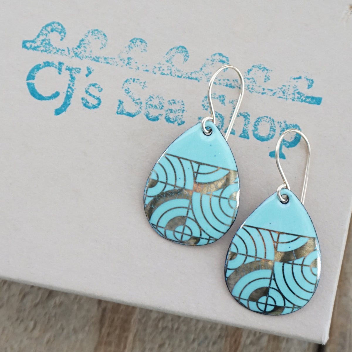Teardrop shaped, seafoam green enamel earrings with gold geometric designs, displayed on a gift box