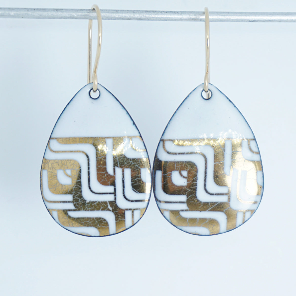 Teardrop shaped, white enamel earrings with gold geometric designs, against a white background