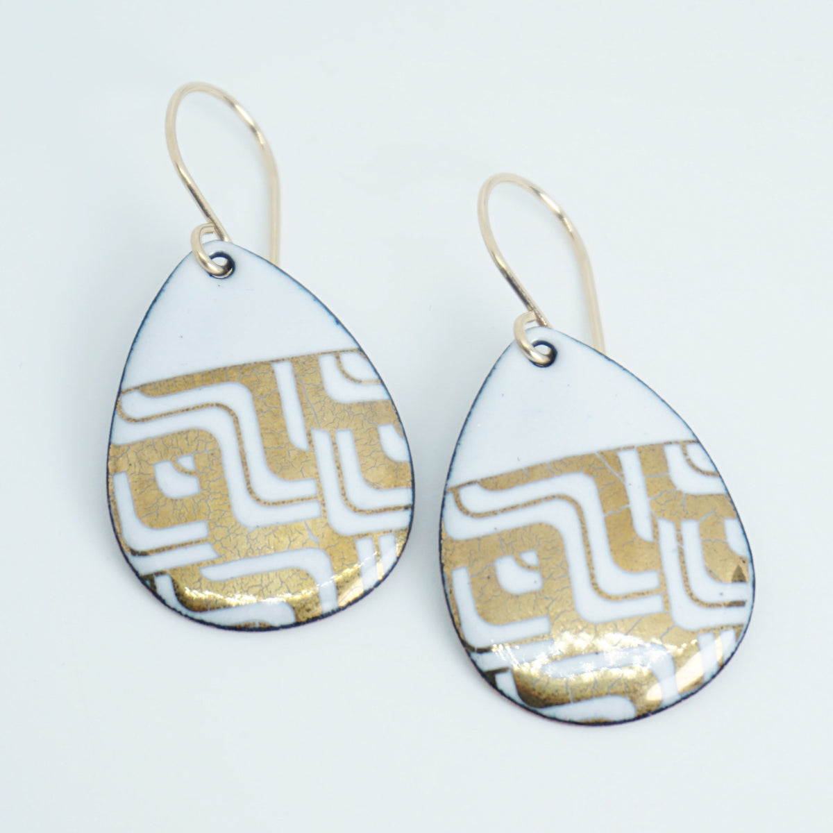 Teardrop shaped, white enamel earrings with gold geometric designs, against a white background