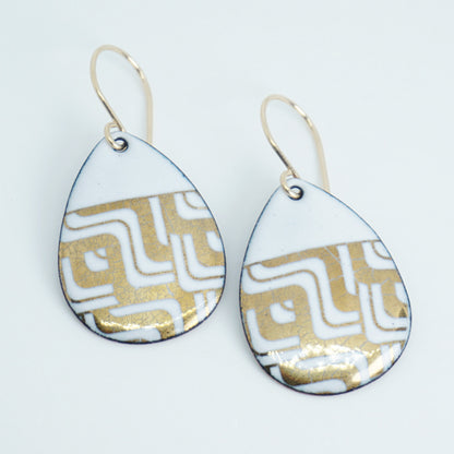 Teardrop shaped, white enamel earrings with gold geometric designs, against a white background