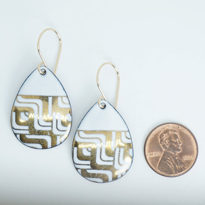 A pair of teardrop-shaped drop earrings with a white and gold geometric maze pattern, shown next to a US penny for scale on a plain white background.