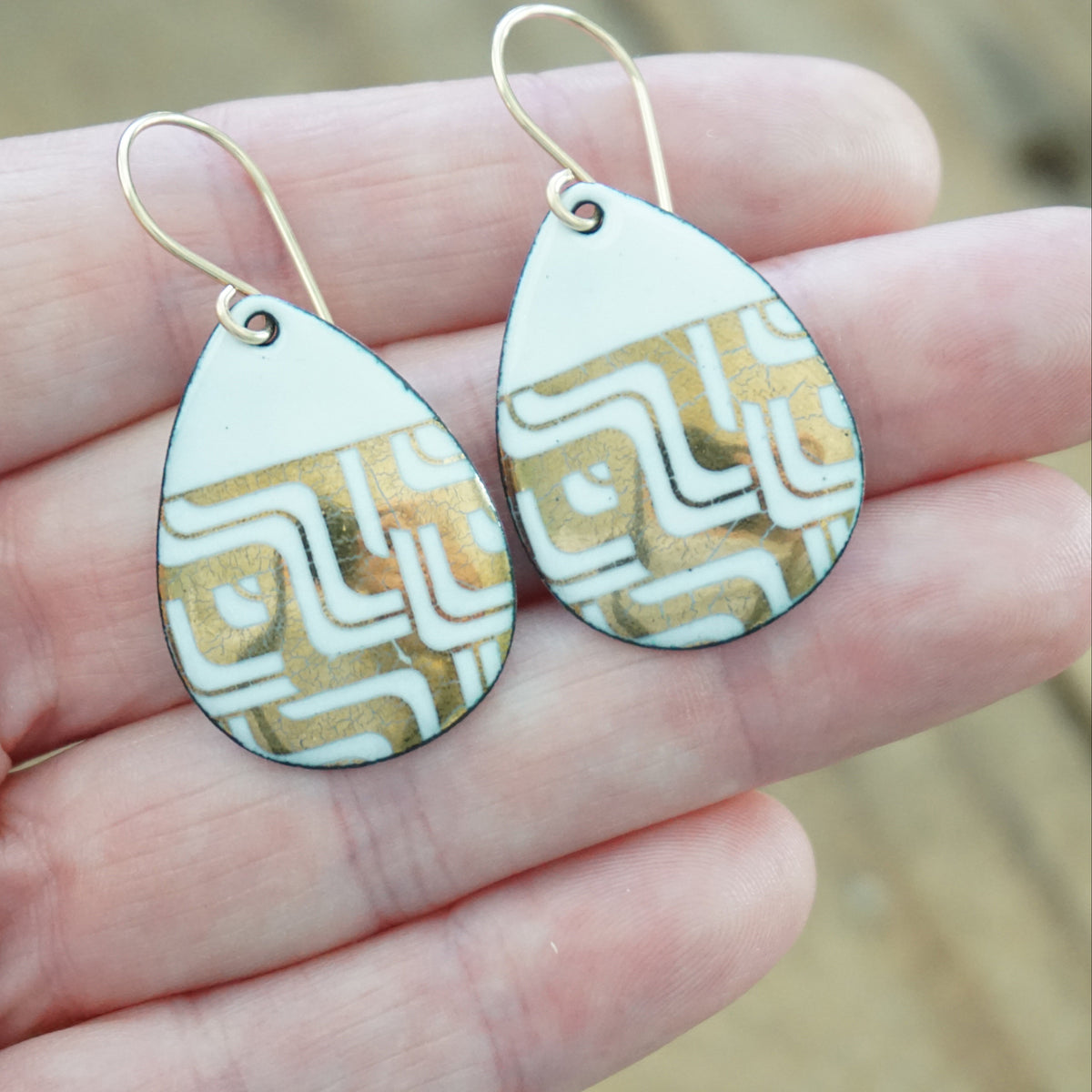 hand holding a pair of Teardrop shaped, white enamel earrings with gold geometric designs, against a wooden background