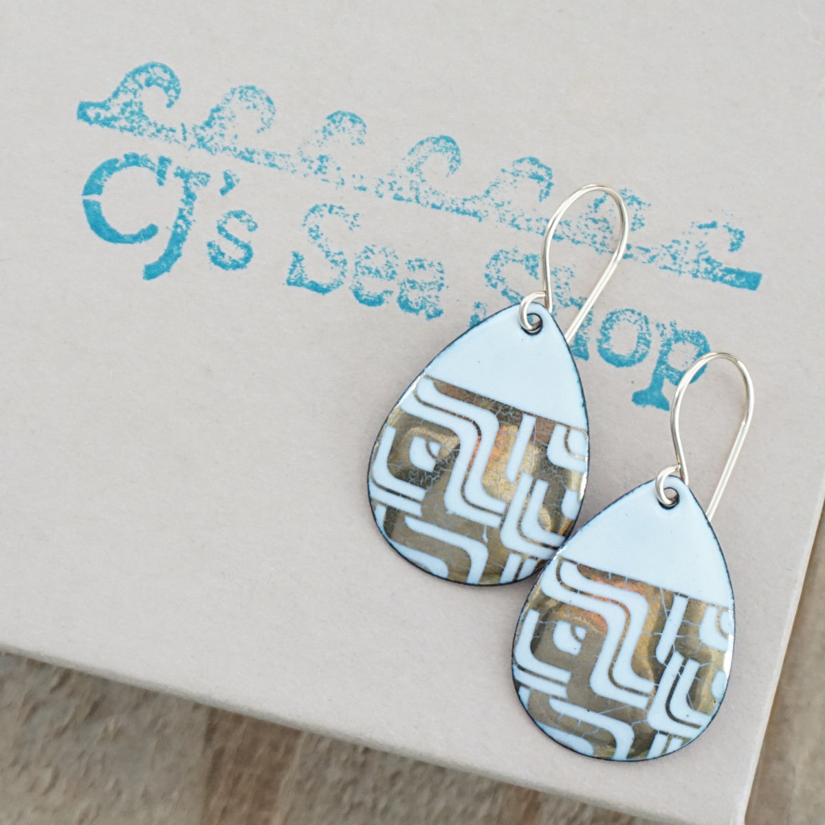 Teardrop shaped, white enamel earrings with gold geometric designs, displayed on a gift box