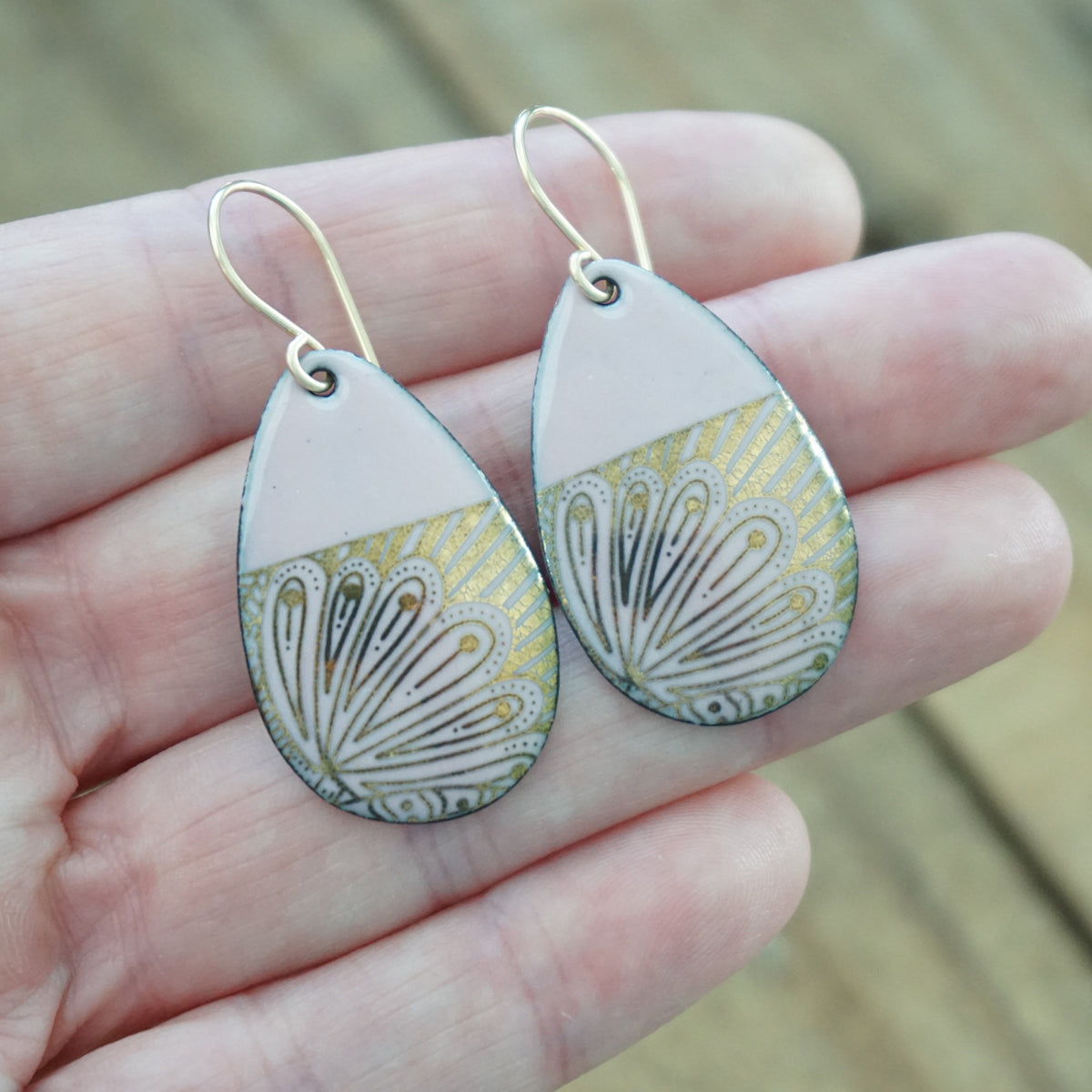 hand holding a pair of Teardrop shaped, pink enamel earrings with gold floral designs, against a wooden background