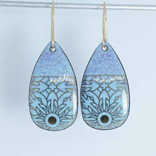 A pair of teardrop-shaped dangling earrings with a light blue and lavender-purple gradient, decorated with gold floral pattern, hanging from gold hooks.