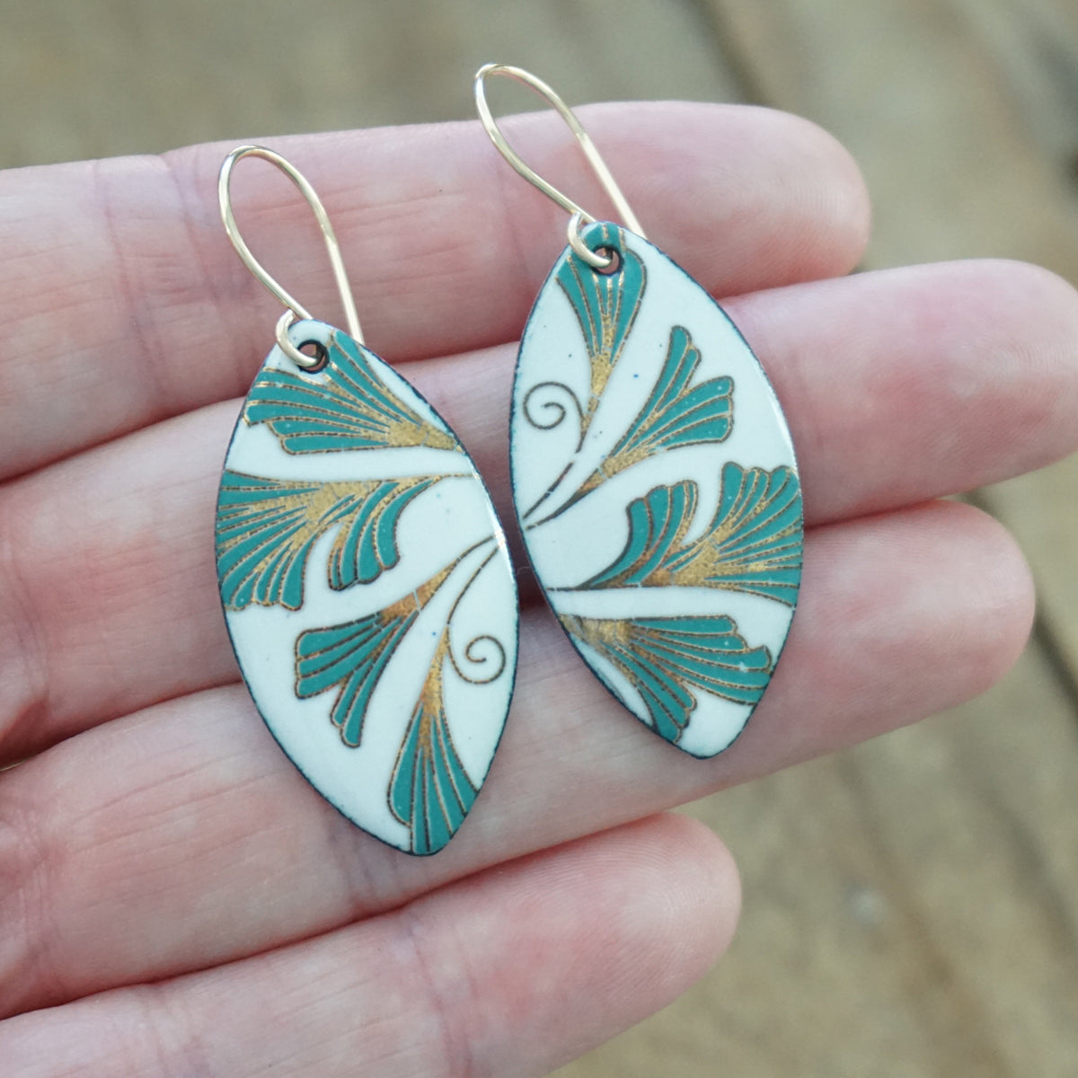 hand holding a pair of Marquis shaped enamel earrings with green and gold leaves, against a wooden background