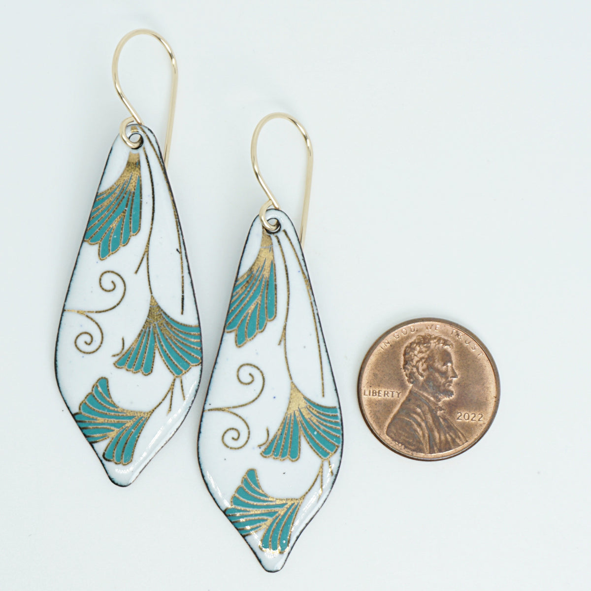 Teardrop shaped enamel earrings with green and gold leaves, next to a penny for scale, against a white background