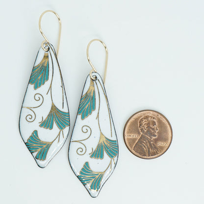 Teardrop shaped enamel earrings with green and gold leaves, next to a penny for scale, against a white background