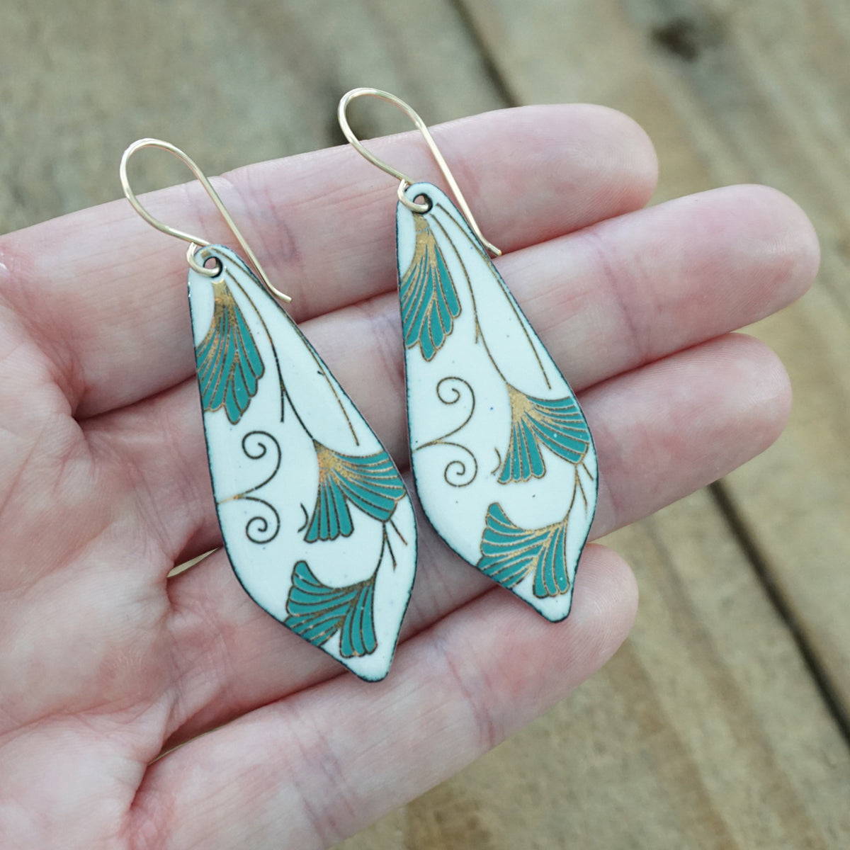 hand holding a pair of Teardrop shaped enamel earrings with green and gold leaves, against a wooden background