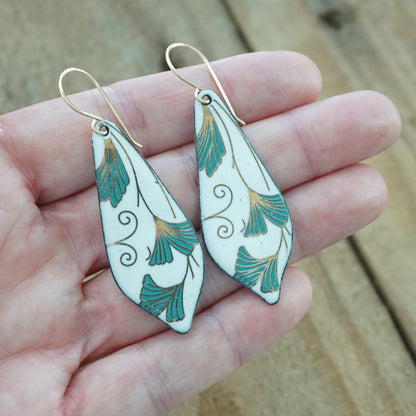 hand holding a pair of Teardrop shaped enamel earrings with green and gold leaves, against a wooden background