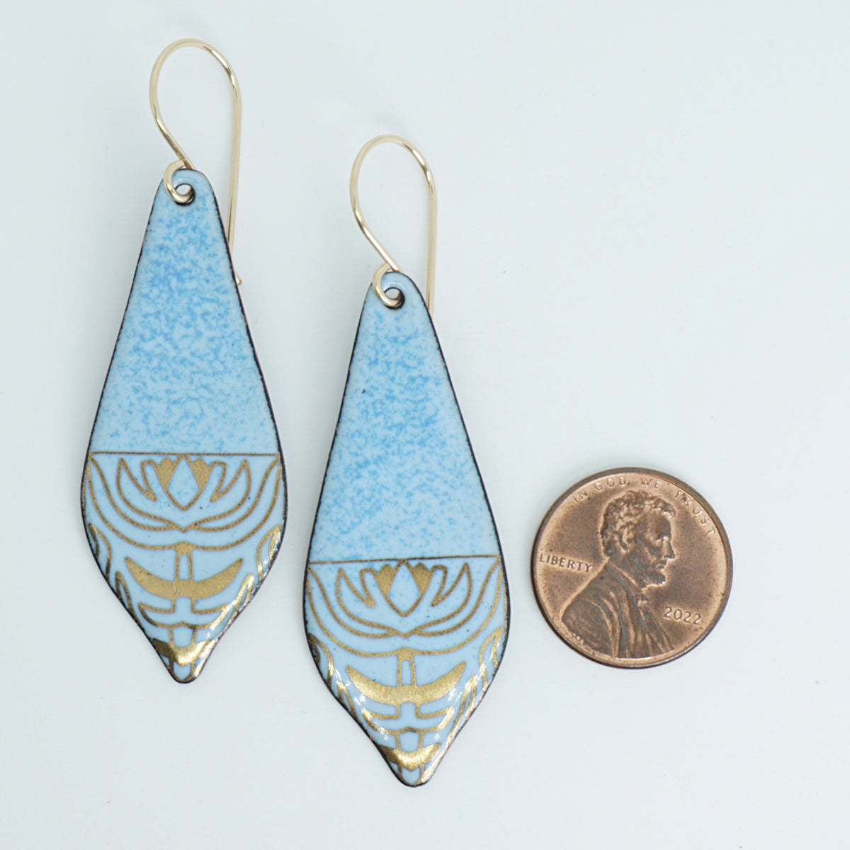 a pair of light blue enamel earrings with gold designs, next to a penny for scale, against a white background