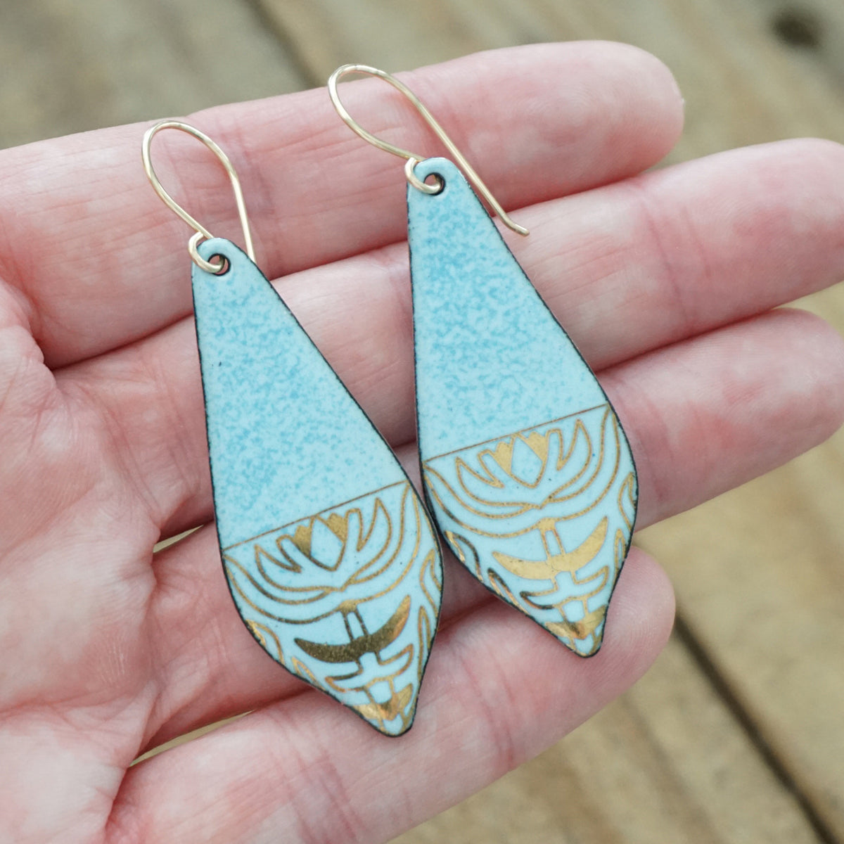 hand holding a pair of light blue enamel earrings with gold designs, against a wooden background