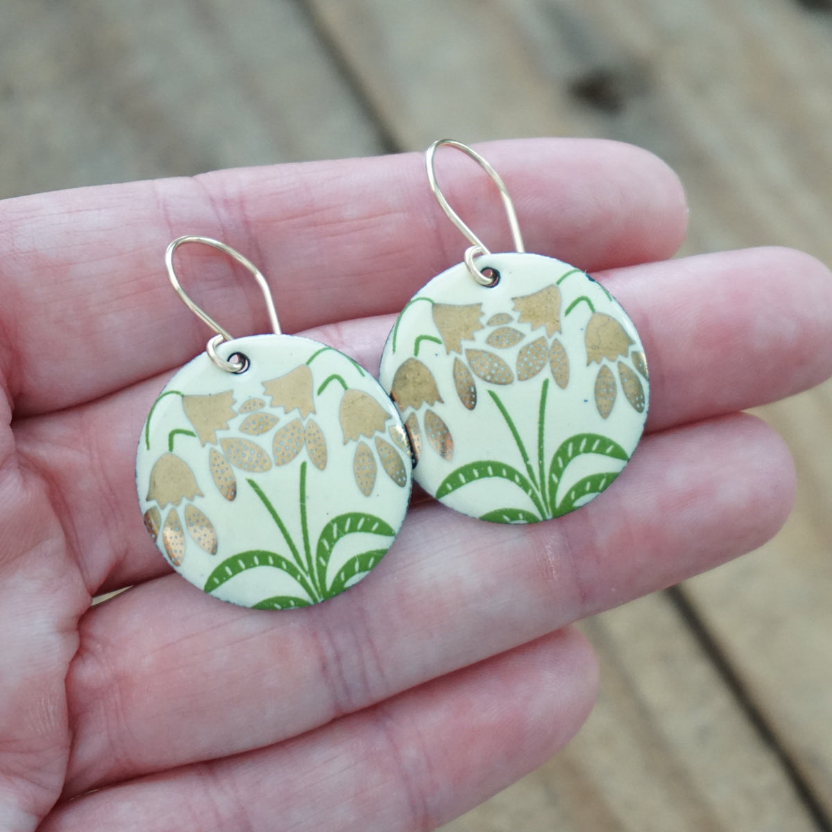hand holding a pair of yellow enamel disc shaped earrings with gold flower accents, against a wooden background