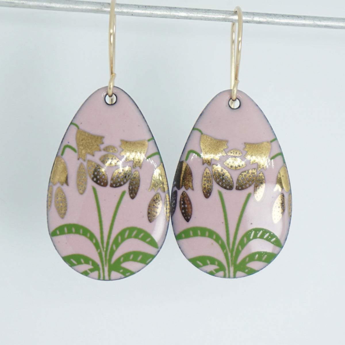 a pair of pink enamel teardrop shaped earrings with gold flower accents, against a white background