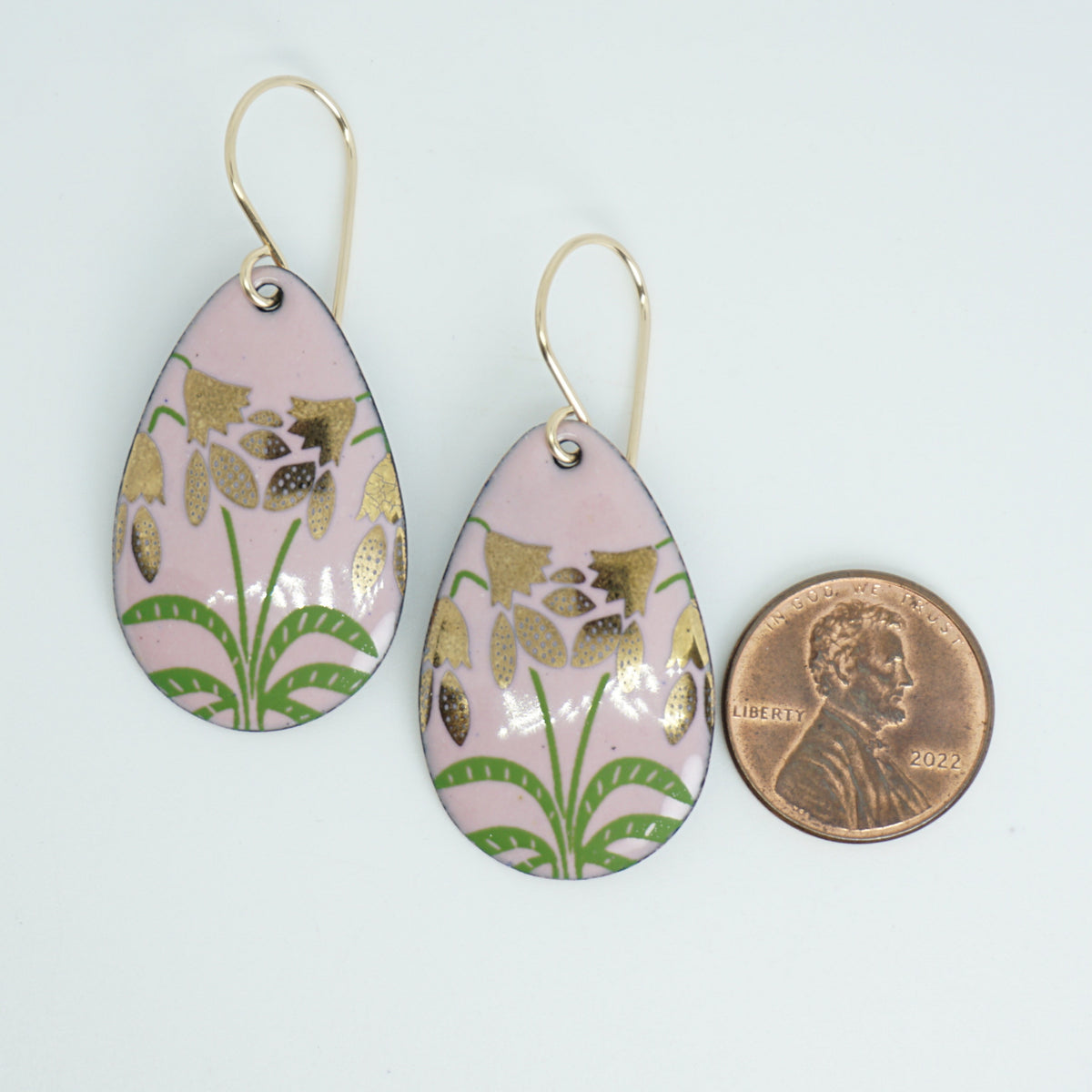 a pair of pink enamel teardrop shaped earrings with gold flower accents, next to a penny for scale, against a white background