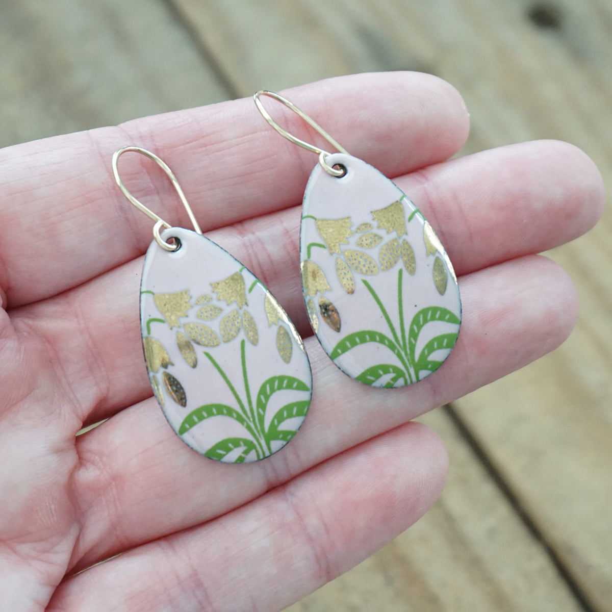 hand holding a pair of pink enamel teardrop shaped earrings with gold flower accents, against a wooden background