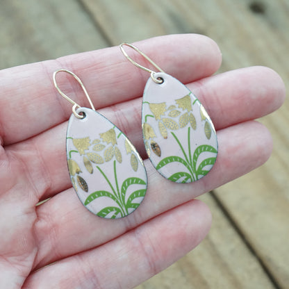 hand holding a pair of pink enamel teardrop shaped earrings with gold flower accents, against a wooden background