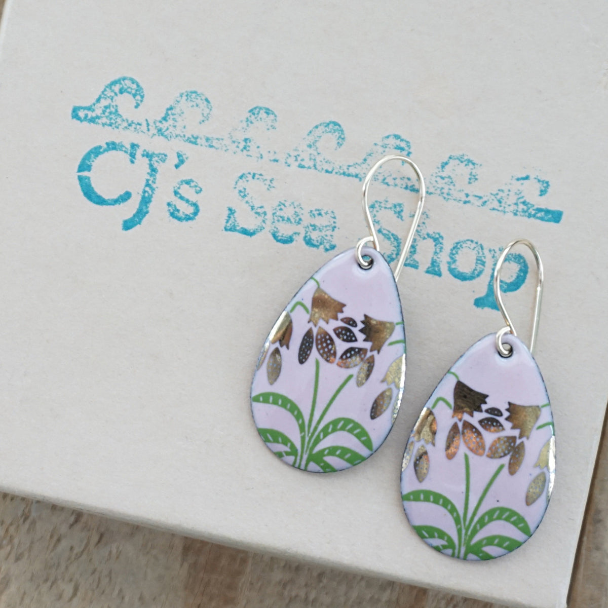 a pair of pink enamel teardrop shaped earrings with gold flower accents, displayed on a gift box