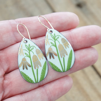 hand holding a pair of seafoam green enamel teardrop shaped earrings with gold flower accents, against a wooden background