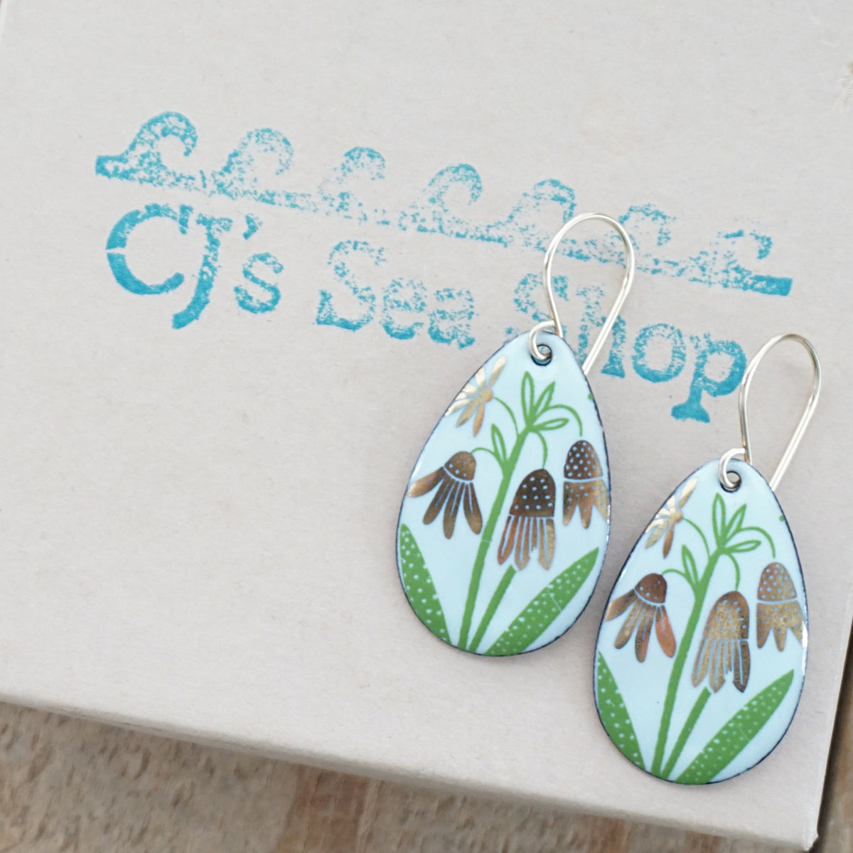 a pair of seafoam green enamel teardrop shaped earrings with gold flower accents, displayed on a gift box