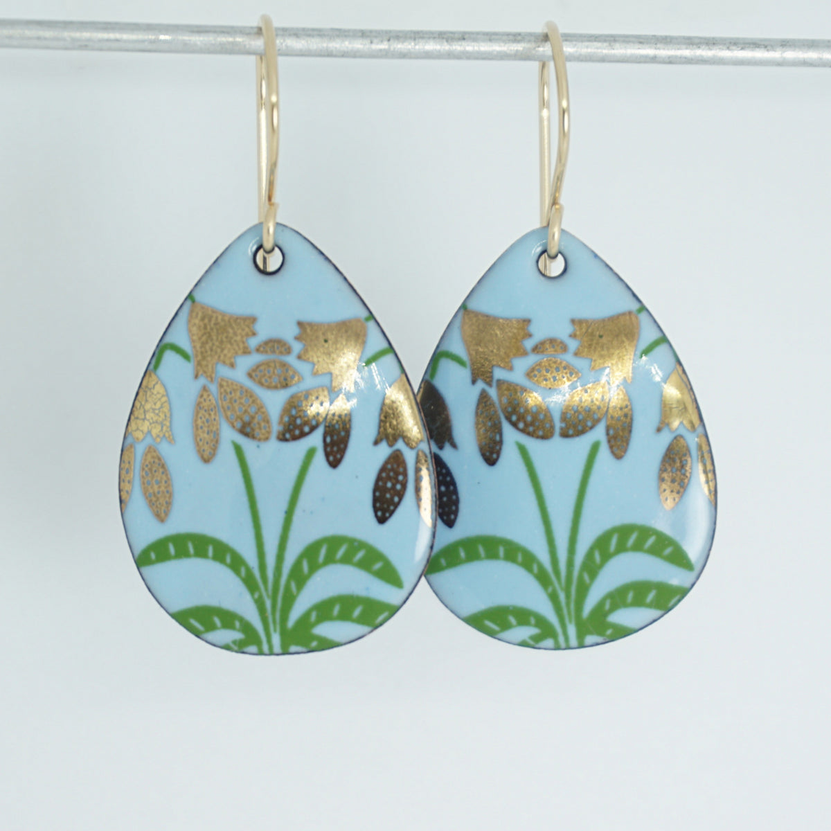 a pair of light blue enamel teardrop shaped earrings with gold flower accents, against a white background