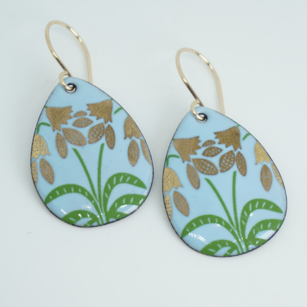 a pair of light blue enamel teardrop shaped earrings with gold flower accents, against a white background