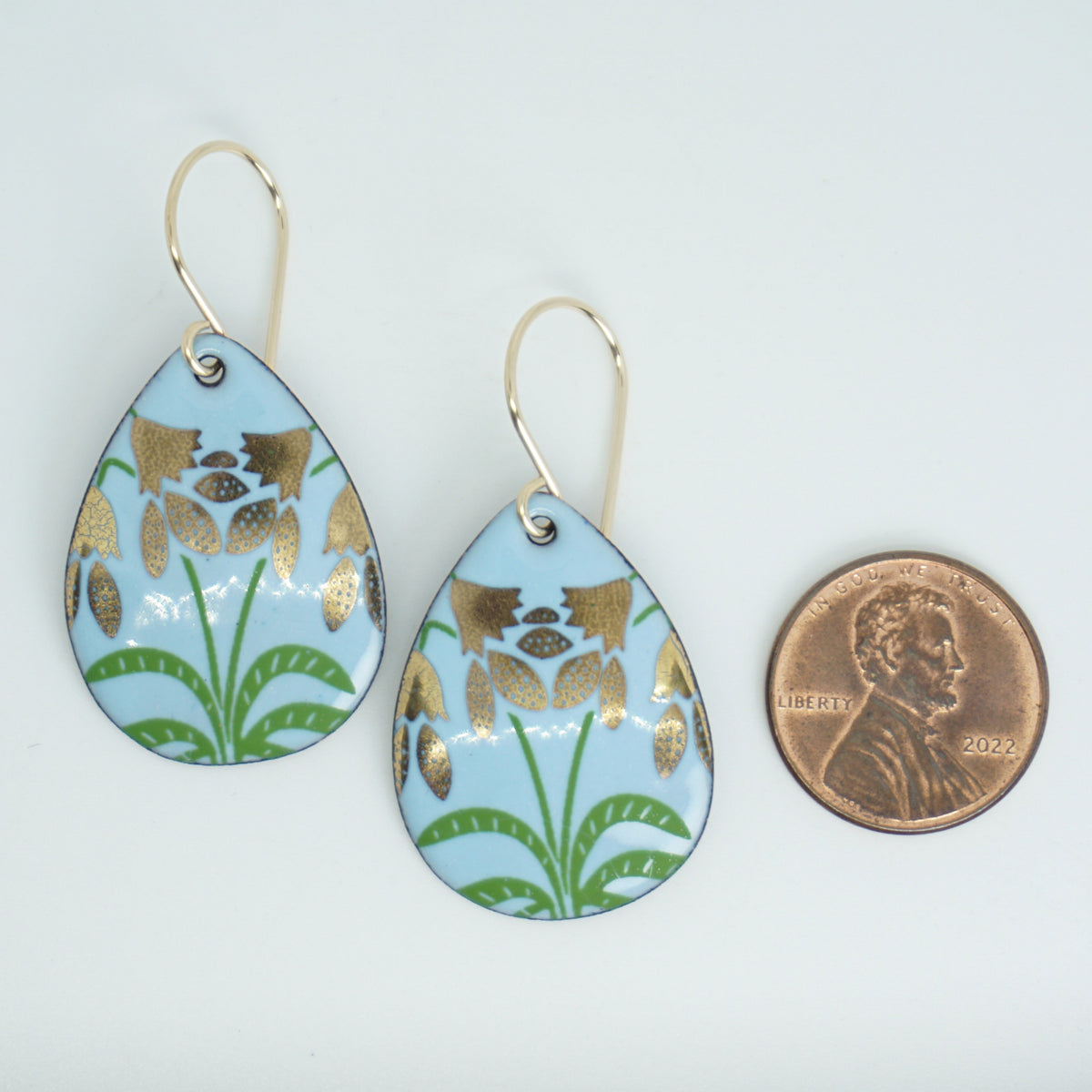 a pair of light blue enamel teardrop shaped earrings with gold flower accents, next to a penny for scale, against a white background