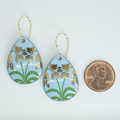 a pair of light blue enamel teardrop shaped earrings with gold flower accents, next to a penny for scale, against a white background