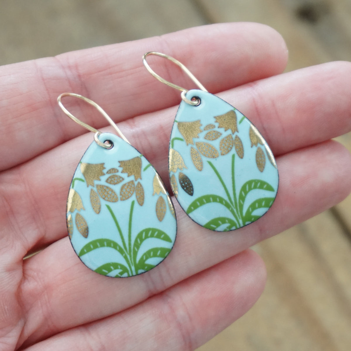 hand holding a pair of light blue enamel teardrop shaped earrings with gold flower accents, against a wooden background