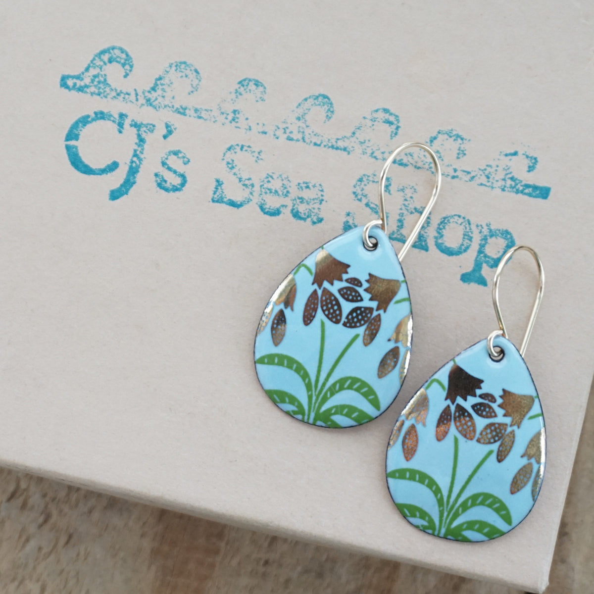 a pair of light blue enamel teardrop shaped earrings with gold flower accents, displayed on a gift box