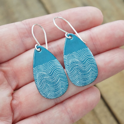 hand holding a pair of teal blue enamel teardrop shaped earrings with white wavy accents, against a wooden background