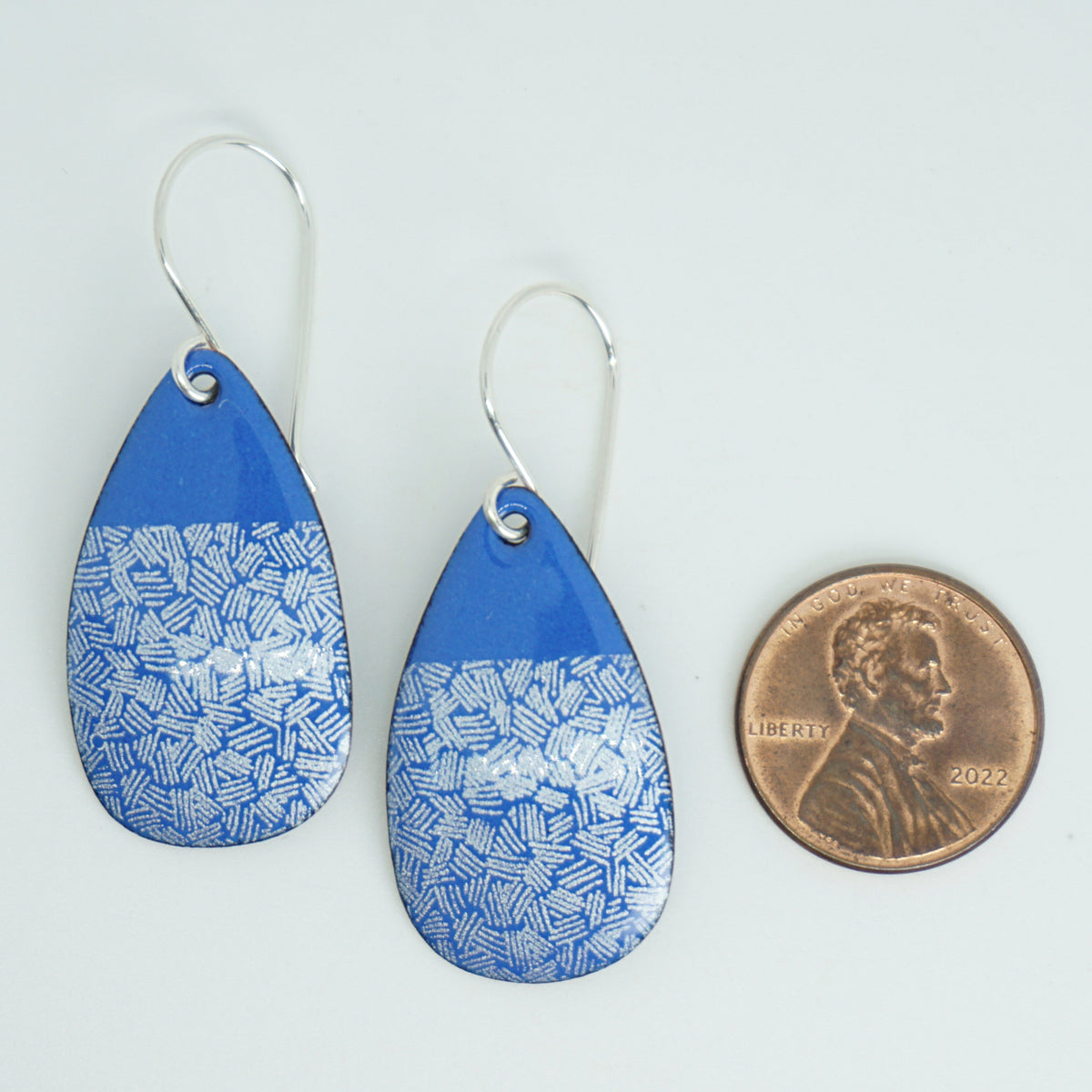 a pair of blue enamel teardrop shaped earrings with white accents, next to a penny for scale, against a white background