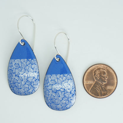 a pair of blue enamel teardrop shaped earrings with white accents, next to a penny for scale, against a white background