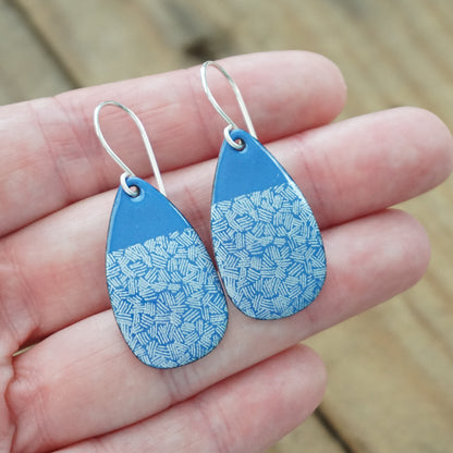 hand holding a pair of blue enamel teardrop shaped earrings with white accents, against a wooden background