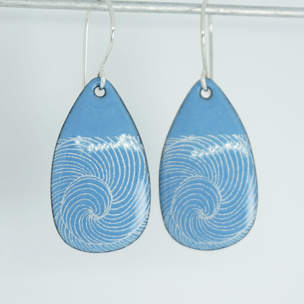 a pair of blue enamel teardrop shaped earrings with white wave accents, against a white background