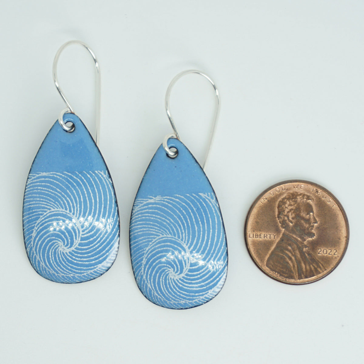 a pair of blue enamel teardrop shaped earrings with white wave accents, next to a penny for scale, against a white background