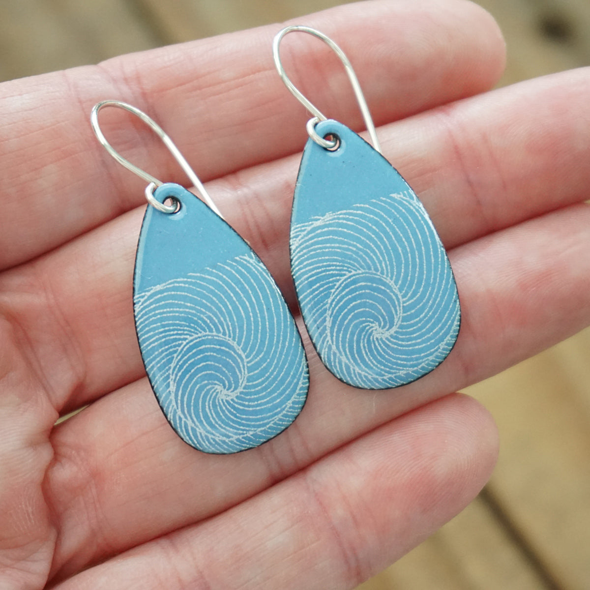 hand holding a pair of blue enamel teardrop shaped earrings with white wave accents, against a wooden background