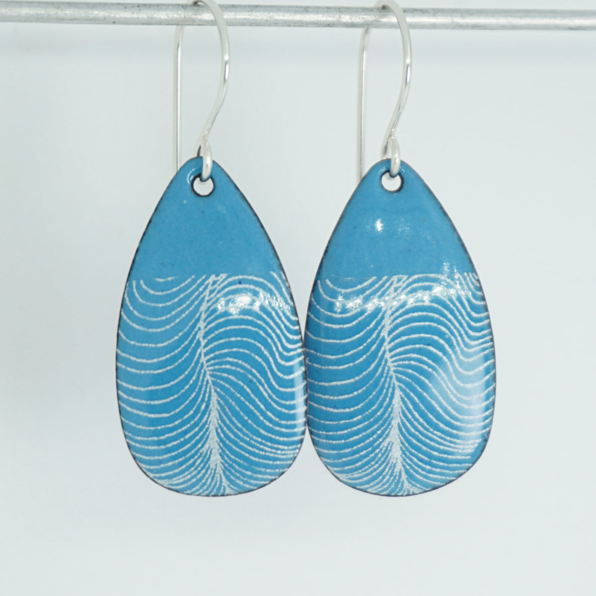a pair of blue enamel teardrop shaped earrings with white wavy accents, against a white background