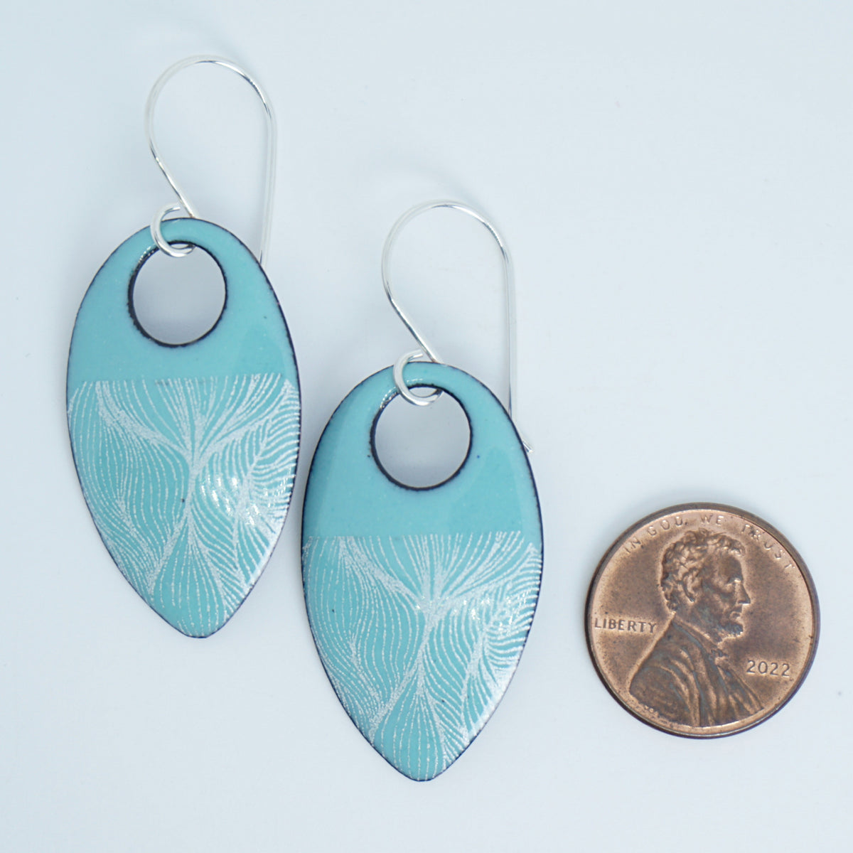 a pair of light green enamel teardrop shaped earrings with white feathery accents, next to a penny for scale, against a white background