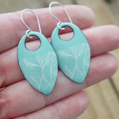 hand holding a pair of light green enamel teardrop shaped earrings with white feathery accents, against a wooden background