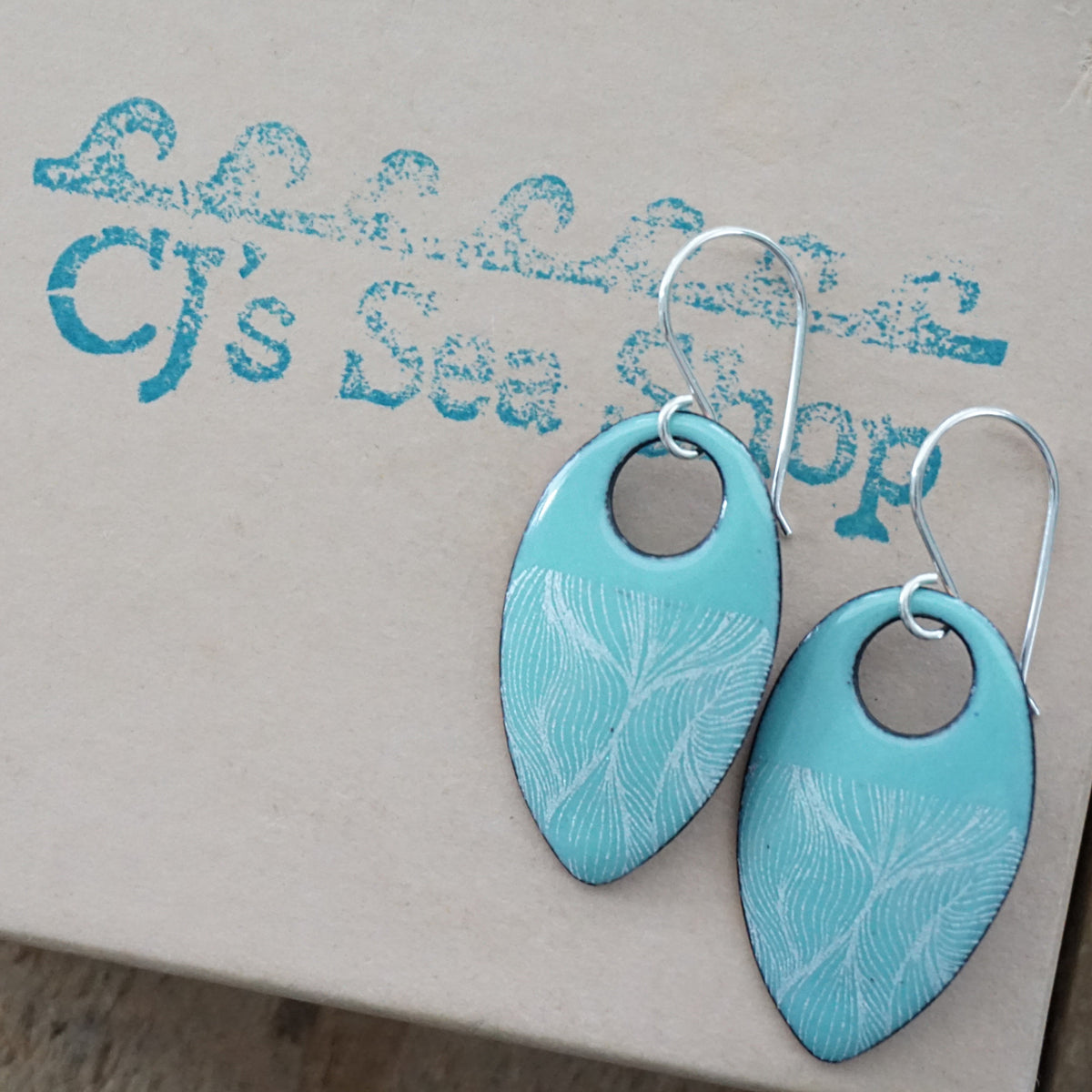 a pair of light green enamel teardrop shaped earrings with white feathery accents, displayed on a gift box