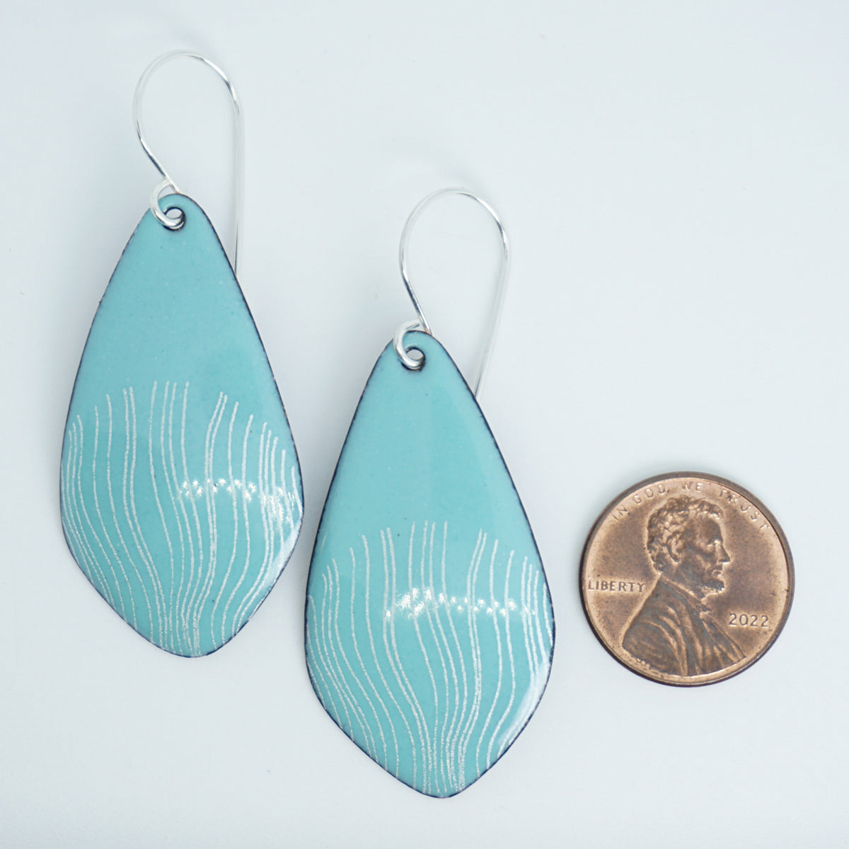 a pair of light green enamel teardrop shaped earrings with white wavy accents,  next to a penny for scale, against a white background