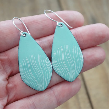 hand holding a pair of light green enamel teardrop shaped earrings with white wavy accents, against a wooden background
