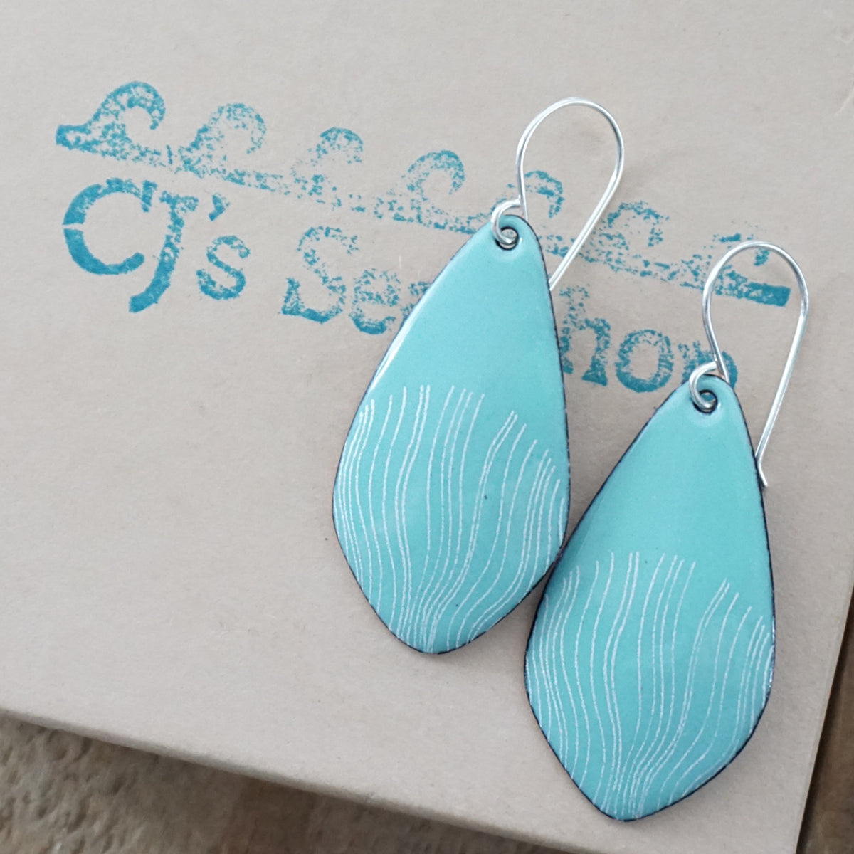 a pair of light green enamel teardrop shaped earrings with white wavy accents, displayed on a gift box