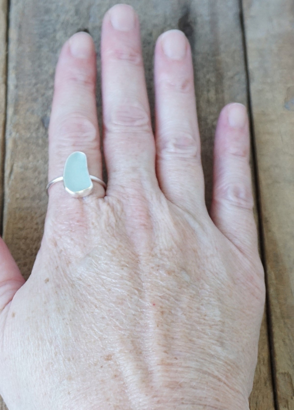 hand wearing a silver ring with a piece of light blue sea glass, against a wooden background