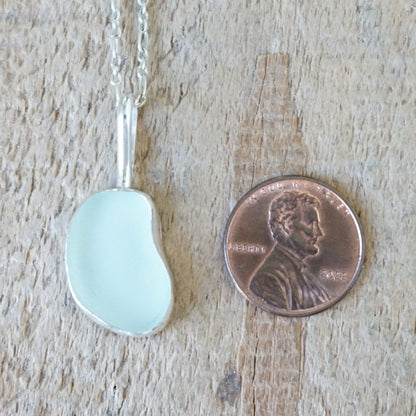 silver pendant with seafoam green sea glass, next to a penny for scale, against a wooden background
