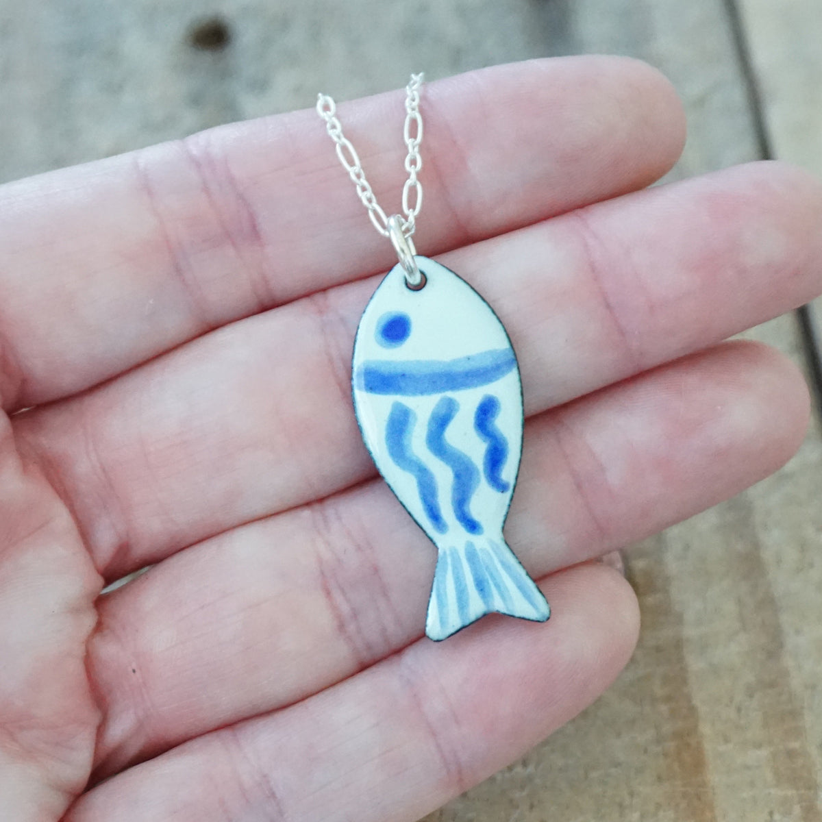 hand holding a light green and blue enamel fish shaped pendant, against a wooden background