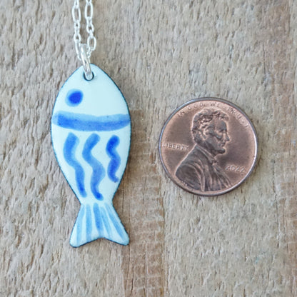 light green and blue enamel fish shaped pendant, next to a penny for scale, against a wooden background