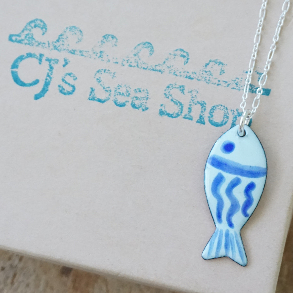 light green and blue enamel fish shaped pendant, displayed on a gift box